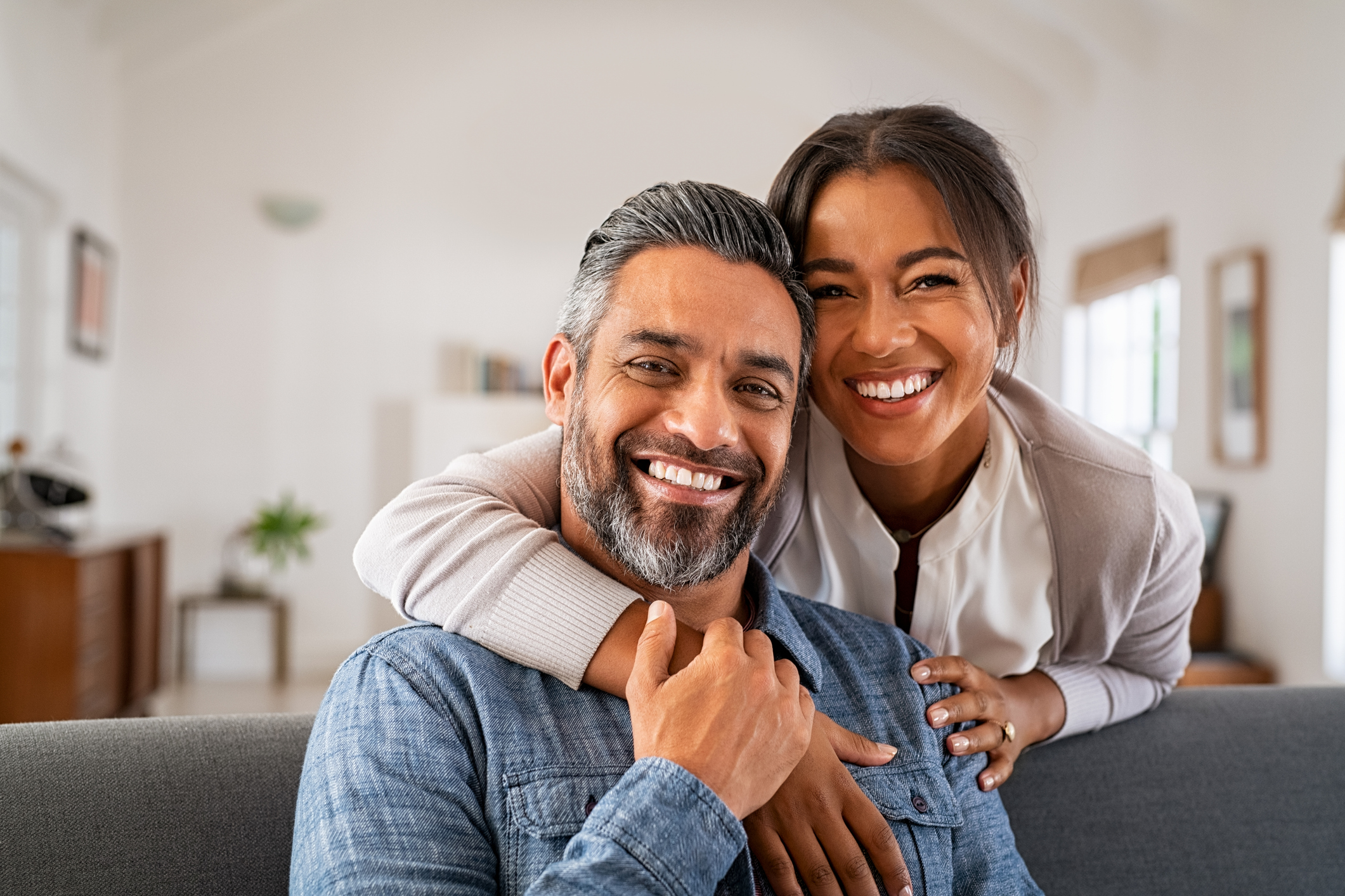 Mature indian couple hugging and looking at camera Portrait of multiethnic couple embracing and looking at camera sitting on sofa. Smiling african american woman hugging mid adult man sitting on couch from behind at home. Happy mature mixed race couple laughing at home.