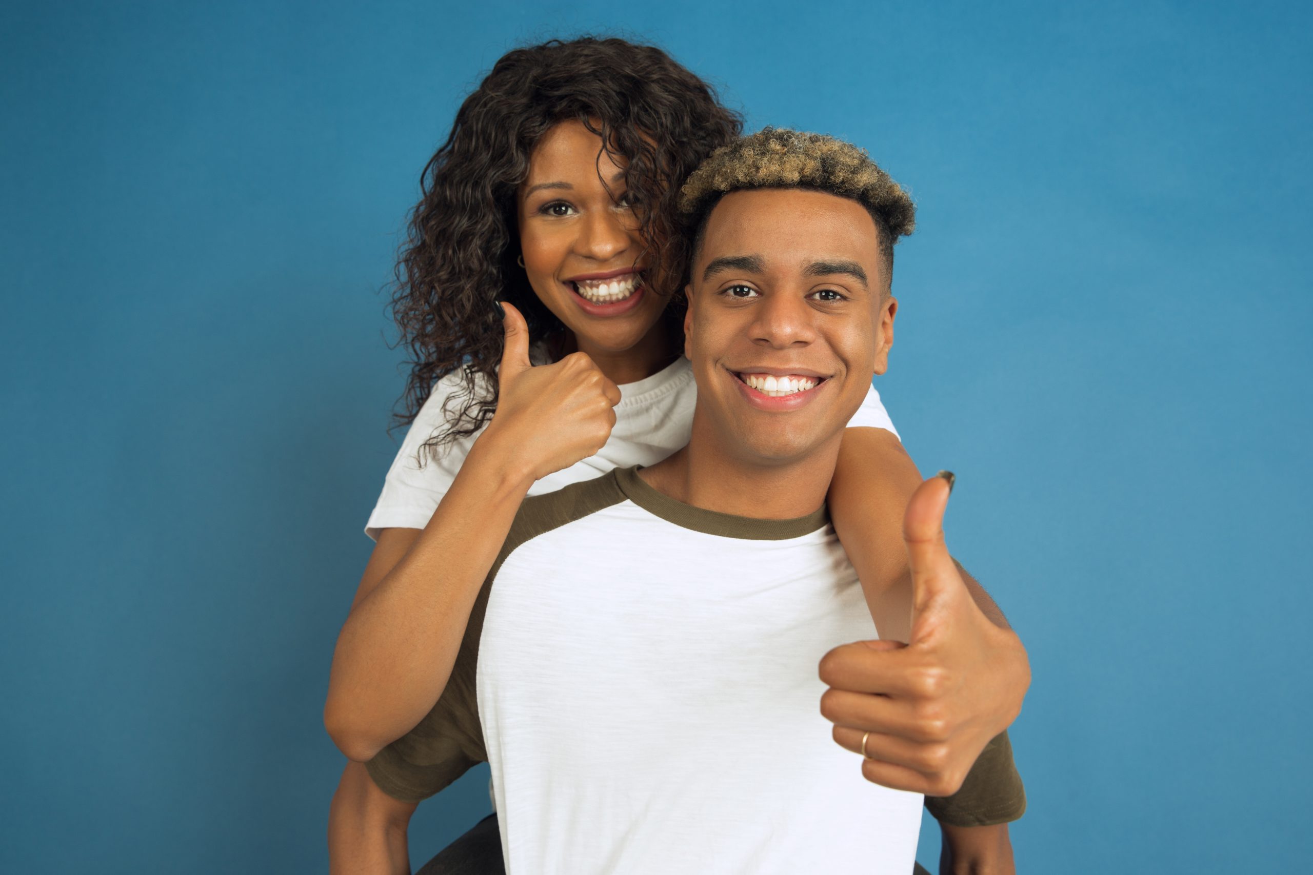 Young emotional african-american man and woman on blue background Young emotional african-american man and woman in white casual clothes posing on blue background. Beautiful couple. Concept of human emotions, facial expession, relations, ad. Hugging, thumbs up.